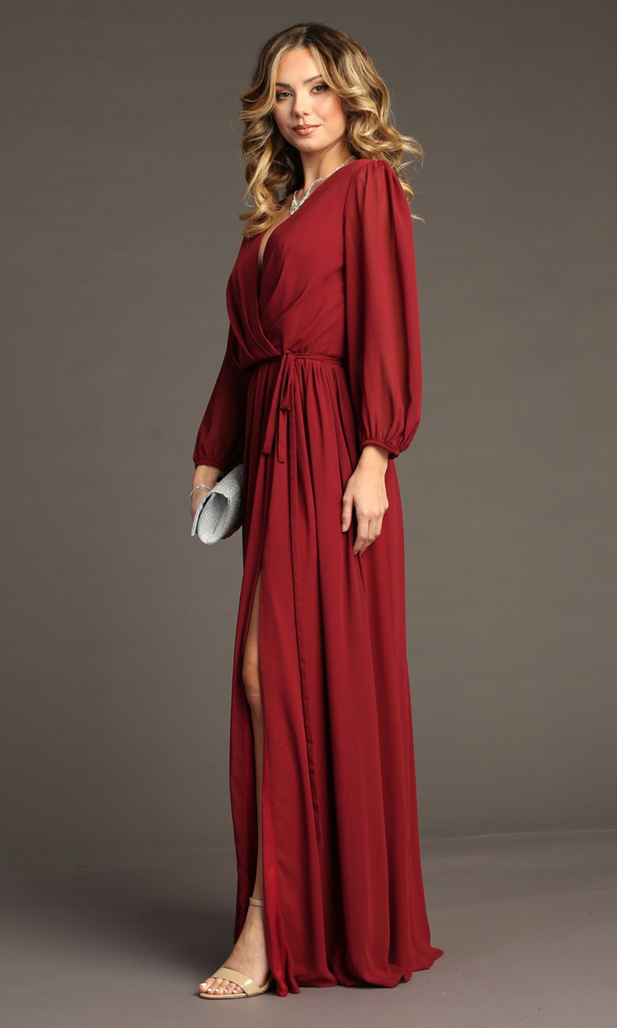 A woman models the Chicas J012B Long-Sleeve Long Formal Dress in maroon, featuring a front slit. She accessorizes with a silver clutch and open-toe heels, posing against a plain background.